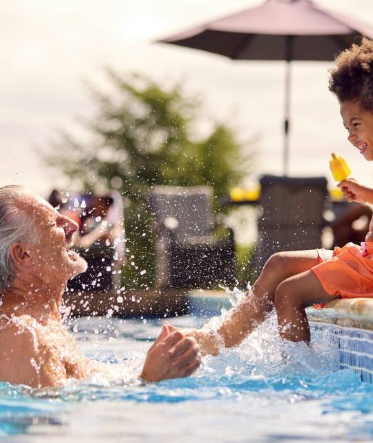 Grandfather And Grandson Eating Ice Lolly At Edge Of Swimming Pool On Summer Holiday